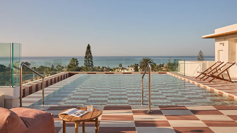A serene rooftop pool with a checkered tile design overlooks the ocean, surrounded by palm trees and a coastal landscape. Lounge chairs and a cozy seating area with a wooden table, an open magazine, and a drink create a relaxing atmosphere. Glass railings offer an unobstructed view of the blue sky and sea, enhancing the tranquil ambiance