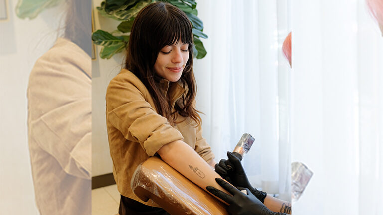 Woman with brown hair and in a brown jacket sits in a chair with her right arm extended as a Tiny Zaps tattoo artist gives her a Tiny Zaps tattoo.