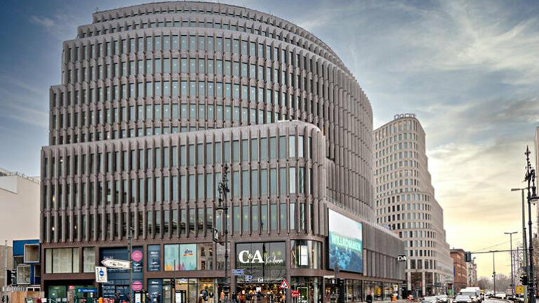 This is a rendering of the exterior of the new Ruby Hotel in Berlin. It's got a neat curved shape and lots of windows, with shops on the bottom floor.  You can see people walking on the sidewalk and lots of cars on the right of the picture waiting at a red light. The sky is a bit cloudy, but you can still see some light. It looks like a busy place where people go to shop and enjoy the hotel whether you’re a guest or local.