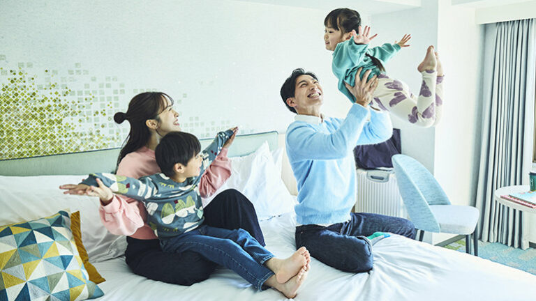 A smiling family sat on a freshly made bed, a 7 year old boy sits on his mother’s lap and the father is throwing a 4 year old girl into the air.